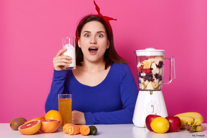 Zaskoczona kobieta trzyma w dłoni szklankę z koktajlem, który przyrządziła w stojącym obok blenderze kielichowym; na blacie leżą owoce